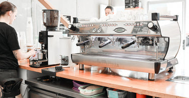 This photo shows a la marzocco coffee machine, Anfim coffee grinder and cinoart automatic tamp sitting on a timber espresso bar bench. There is a female barista wearing black taking a coffee order from a customer.
