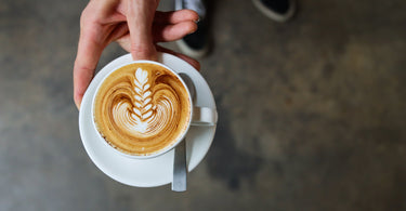 Picture of a hand holding out a cappuccino on in a white cup and saucer. 