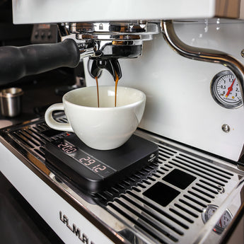 A shot of espresso from a white La Marzocco Linea Mini coffee machine dropping into a white porcelain cup which is sitting on a set of Acaia Lunar coffee scales to measure the extracted espresso. 